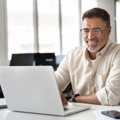 Happy smiling mature Indian or Latin business man ceo trader using computer, typing, working in modern office, doing online data market analysis, thinking planning tech strategy looking at laptop.